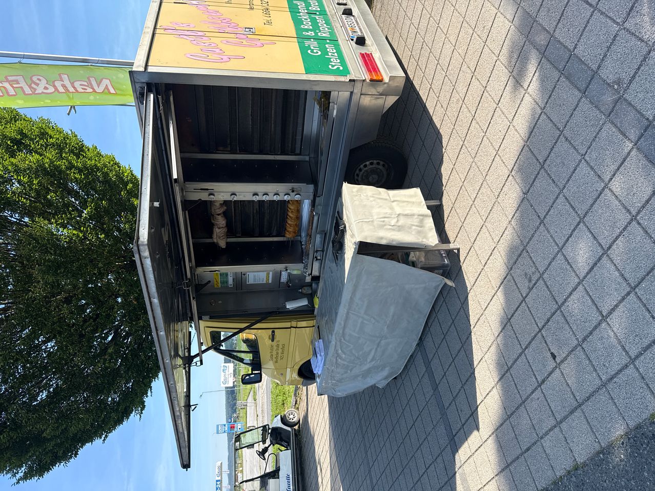 Ein Foodtruck mit offener Heckklappe, am Straßenrand geparkt. Der Truck hat ein grünes Schild mit der Aufschrift 'Grill & Backofen'. Ein grauer Stoff bedeckt einen Tisch davor.