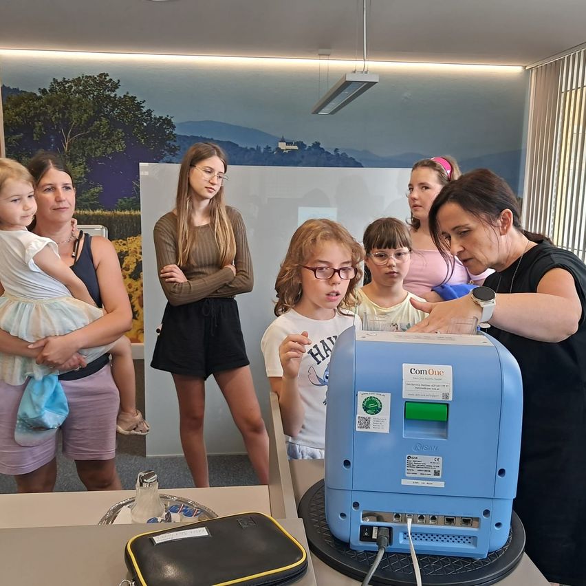 Eine erwachsene Frau steht mit einem Kind und anderen Kindern und beobachtet alle ein blaues Gerät. Die Erwachsene trägt eine Uhr, und die Kinder tragen eine Brille. Das Gerät hat einen Aufkleber mit der Aufschrift Com One.