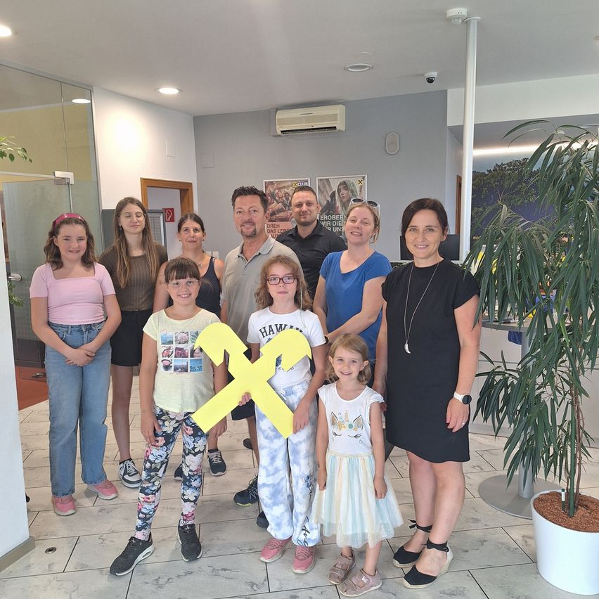 Eine Gruppe von Menschen steht in einem Büro. Einige lächeln, während andere in die Kamera schauen. Ein Mädchen hält einen gelben Buchstaben X. Ein Topf mit einer Pflanze steht rechts.