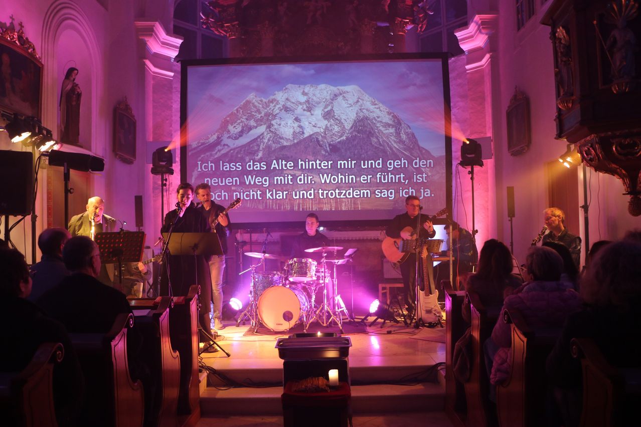 Eine Band tritt auf einer Bühne auf, mit einer Projektionsleinwand im Hintergrund, die einen Berg zeigt. Auf der Leinwand steht 'Ich lass das Alte hinter mir und geh den neuen Weg mit dir.' Das Publikum sitzt davor.