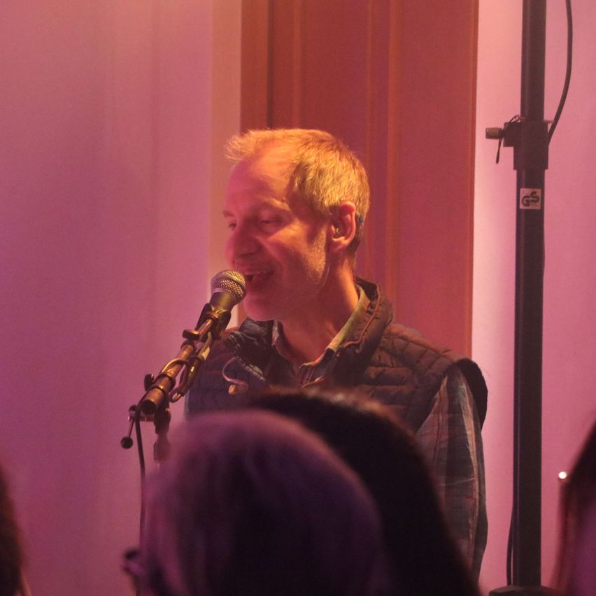 Ein Mann in einem karierten Hemd und Weste singt in ein Mikrofon in einem schwach beleuchteten Raum mit einer rosa Wand.