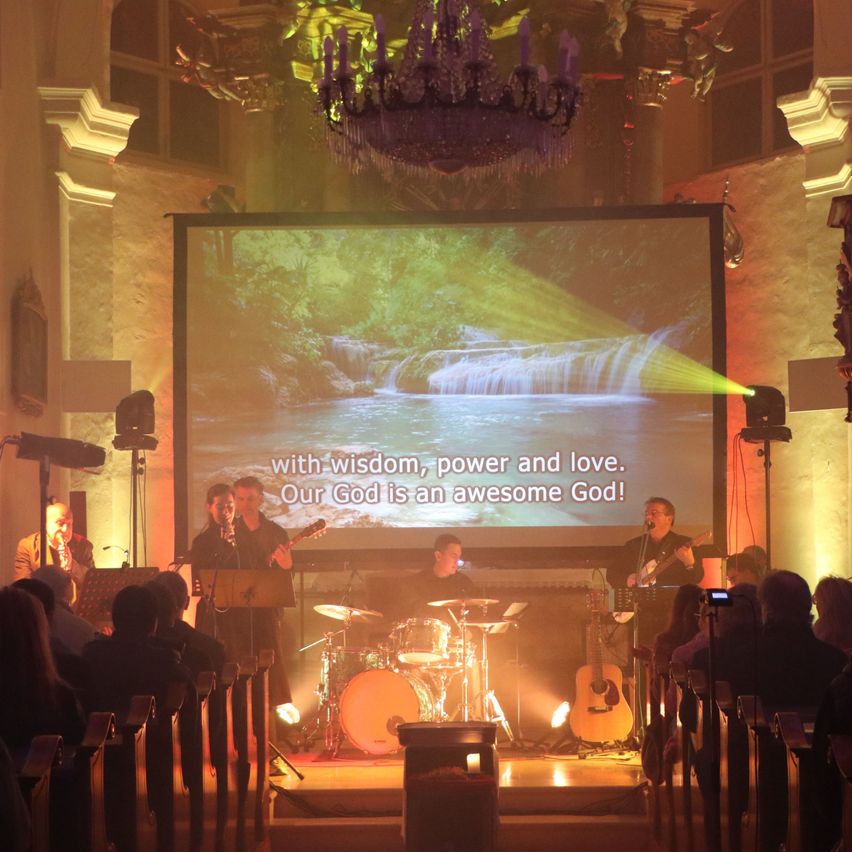 Eine Band spielt auf der Bühne mit einer Leinwand im Hintergrund, auf der ein Wasserfall zu sehen ist. Der Text auf der Leinwand lautet: 'Mit Weisheit, Kraft und Liebe. Unser Gott ist ein großartiger Gott!'