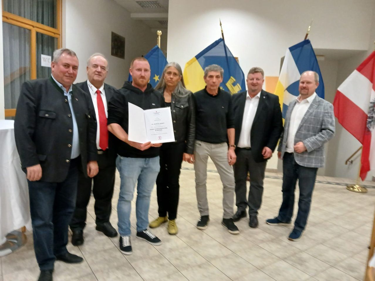 Eine Gruppe Erwachsener steht drinnen. Ein Mann hält ein Zertifikat und sie alle tragen formelle Kleidung. Flaggen sind hinter ihnen.