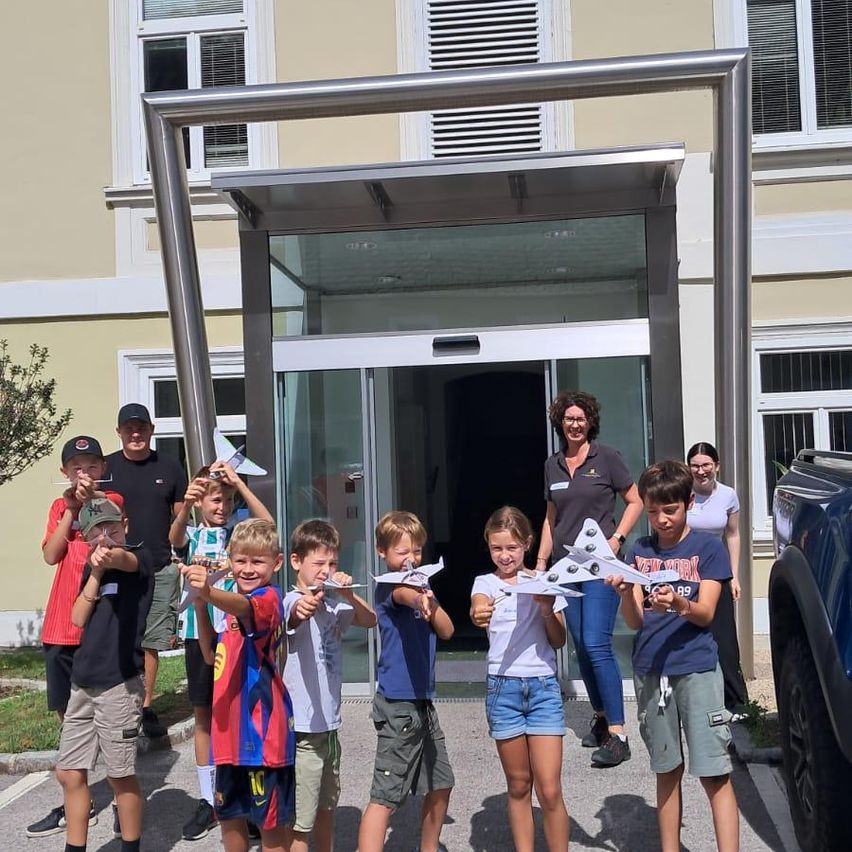 Eine Gruppe von Kindern und zwei Erwachsenen steht vor einem Gebäude und hält Papierflugzeuge in der Hand. Die Kinder lächeln und scheinen für ein Foto zu posieren.
