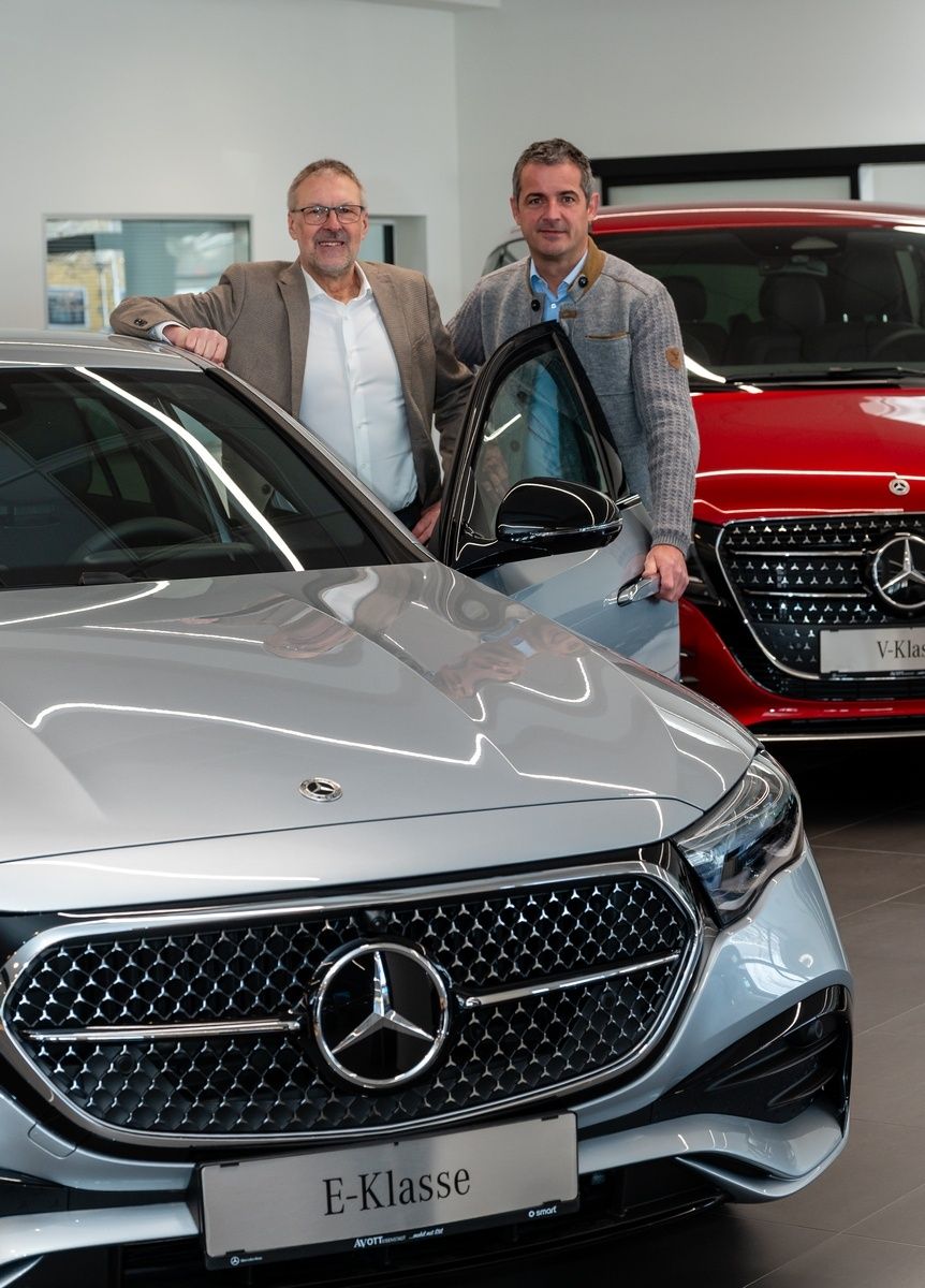 Zwei Männer stehen neben einem silbernen Mercedes-Benz, ein roter Wagen dahinter. Ein Mann berührt das silberne Auto, der andere lächelt.