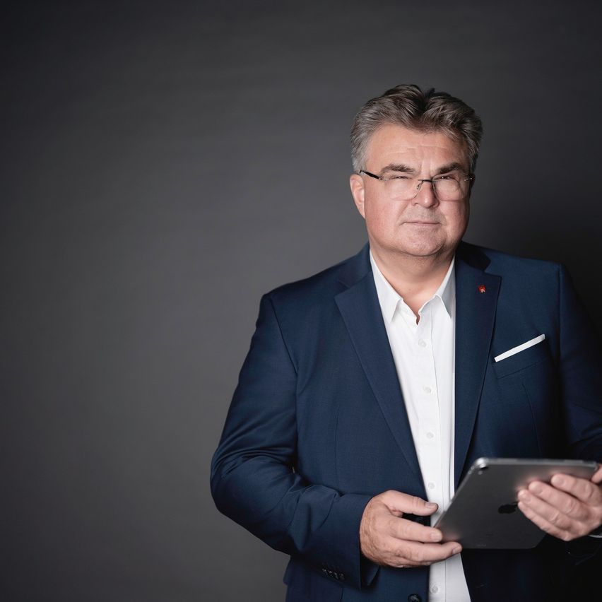 Ein Mann in einem blauen Anzug und Brille hält ein Tablet und steht vor einem grauen Hintergrund.