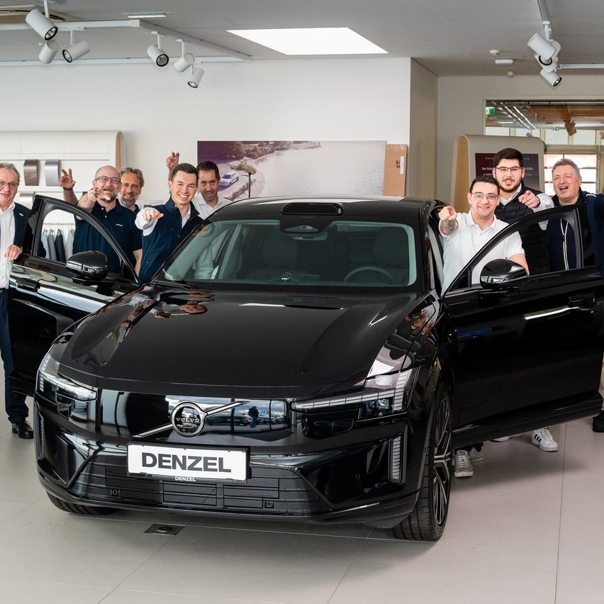 Eine Gruppe von Menschen steht in einem Showroom, neben einem schwarzen Volvo-Auto mit dem Kennzeichen 'DENZEL'. Die Türen des Autos sind geöffnet, und sie wirken aufgeregt.