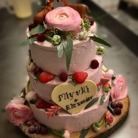 Ein dreistöckiger rosa Kuchen, verziert mit rosa Blumen, grünen Blättern und Erdbeeren. Es zeigt ein Herz mit dem Text 'Miyu + Alexander'. Oben sitzen Rehenfiguren.