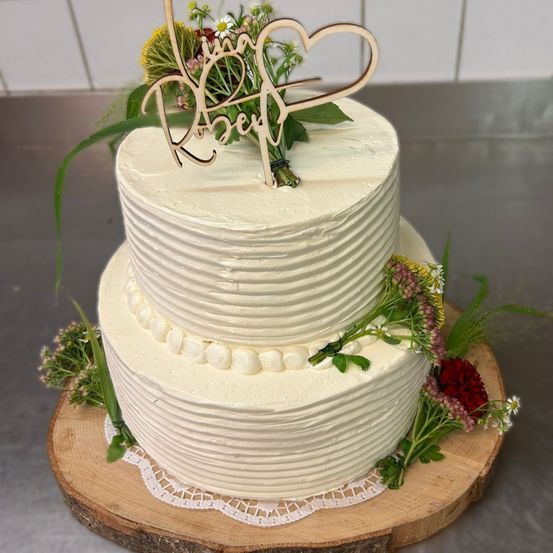 Ein dreistufiger weißer Kuchen mit weißer Glasur und weißen Perlen an den Seiten. Oben ist ein Holzherzzeichen und Blumen. Der Kuchen steht auf einem Holzbrett mit Spitzenbordüre.
