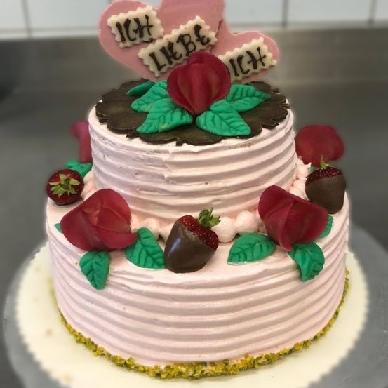 Ein rosa Kuchen mit drei Schichten. Jede Schicht ist mit Frosting, Schokoladenblättern und roten Rosen verziert. Die Spitze ist mit einem Herz und dem Wort 'ICH LIEB' verziert.