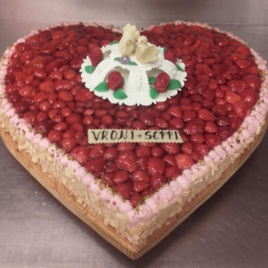 Ein herzförmiger Kuchen, dekoriert mit roten Erdbeeren, weißer Creme und einem Paar Hasen obenauf, mit dem Text Vroni+Seppi.