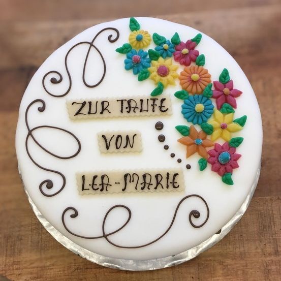Ein Kuchen mit einer weißen Zuckergussbasis, dekoriert mit bunten Blumen und den Worten 'ZUR TAUFE VON LEA-MARIE' in Braun geschrieben.
