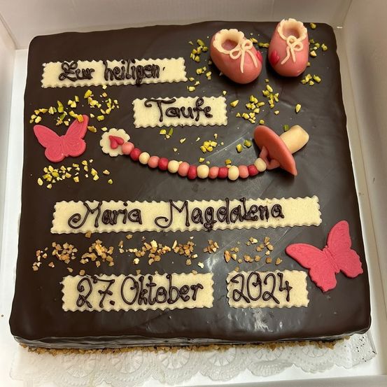 Ein Schokoladenkuchen mit Schokoladenglasur, verziert mit Babyschuhen, einem Schnuller und Schmetterlingen. Es steht 'Maria Magdalena' und das Datum '27. Oktober 2024'.