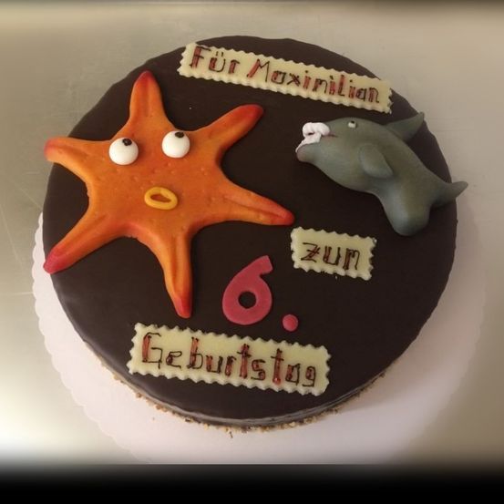 Ein Schokoladen-Geburtstagskuchen mit einem Seestern und einem Haifisch darauf. Es steht 'Für Maximilian', 'zum', '6.', und 'Geburtstag'.