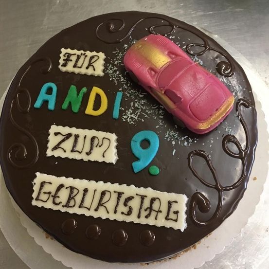 Ein Schokoladenkuchen mit Zuckerguss verziert und einer rosafarbenen Autofigur oben. Es steht 'Für Andi Zum 9. Geburtstag' in bunten Buchstaben.