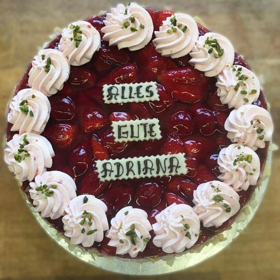 Ein Kuchen mit roter Erdbeerfüllung, oben mit Schlagsahne und weißer Glasur. Es hat weißen Schriftzug, der 'ALLES GUETE ADRIANA' darauf steht.