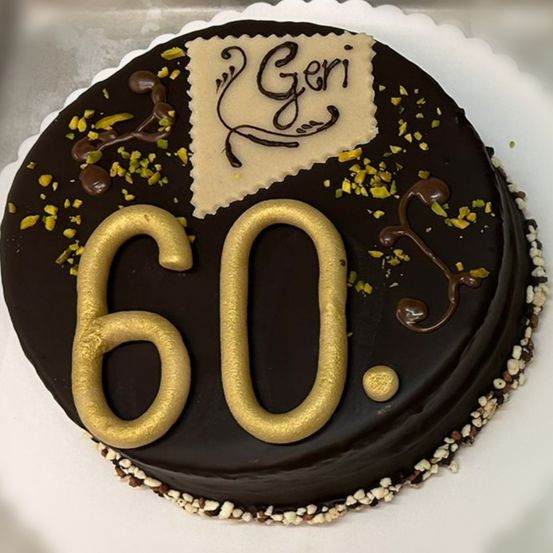 Ein Schokoladenkuchen mit der Nummer 60 in Gold, einer weißen Karte mit Geri in Schwarz und Schokolade und Pistazien oben.