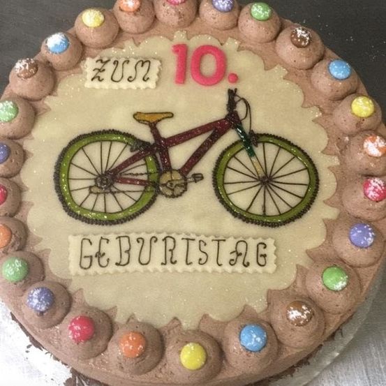 Ein Schokoladenkuchen mit Fahrraddesign und Geburtstagsbotschaft. Der Kuchen hat bunte Bonbons am Rand.