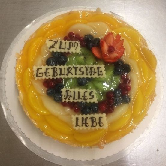 Ein Obstkuchen mit verschiedenen Früchten und Glasur dekoriert, obenauf eine Blume. Der Kuchen hat eine in deutscher Sprache geschriebene Nachricht, die 'Zum Geburtstag Alles Liebe' bedeutet.