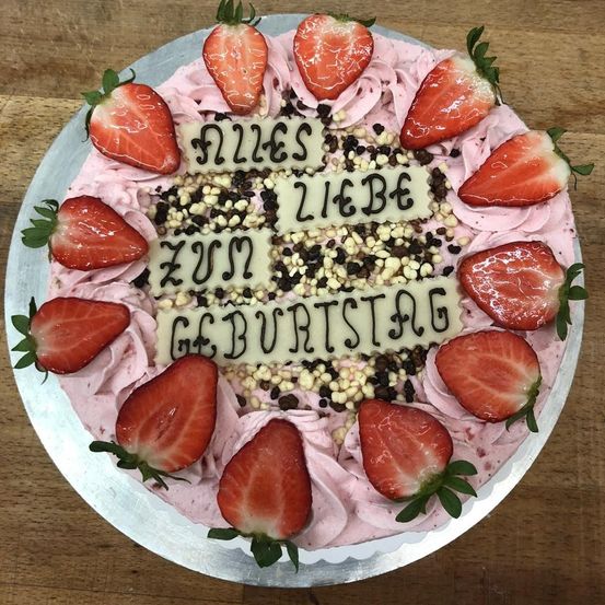 Ein rosa Kuchen mit weißer Glasur ist mit Erdbeeren und schwarzen und weißen Streuseln verziert. Der Kuchen sagt 'Happy Birthday' in weißer Glasur.