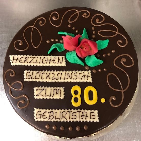 Ein Schokoladenkuchen mit 'Herzlichen Glückwunsch zum 80. Geburtstag' darauf, mit zwei roten Rosen und Schokoladenverzierungen.