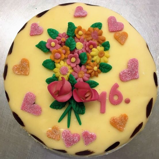 Ein runder Geburtstagskuchen mit gelber Basis, dekoriert mit pinken und gelben Blumen, Herzen und der Nummer 16 in Pink geschrieben.