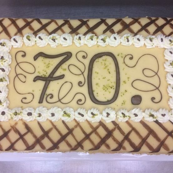 Ein großer quadratischer Kuchen mit '70' in Braun auf cremefarbenem Hintergrund, umgeben von weißer Glasur und grünen Dekorationen.