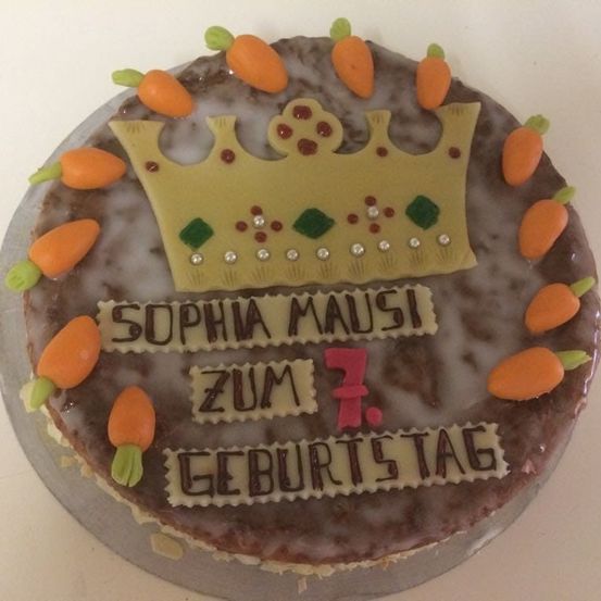 Ein brauner Kuchen mit einer goldenen Krone und Karottendekorationen. Es steht 'Sophia Mausi zum 7. Geburtstag'. Der Kuchen steht auf einem grauen Teller.