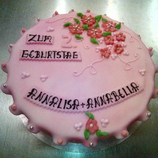 Ein rosa Kuchen mit dekorativen Blumen und den Worten Zum Geburtstag und Annalisa+Annabella darauf. Der Kuchen ist rund mit rosa Glasur.