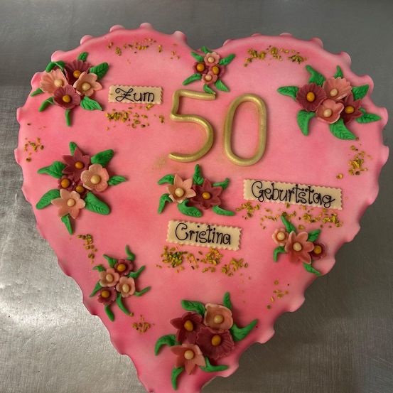 Ein rosa Herzförmiger Kuchen, dekoriert mit Blumen, goldener Zahl 50 und den Namen Zum, Geburtstag und Cristina.