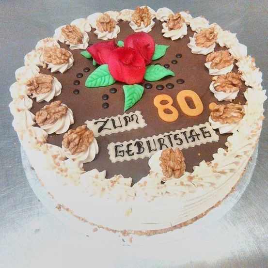 Ein Schokoladenkuchen mit Walnüssen, einer roten Rose und einem Schokoladenrand. Der Kuchen steht für ZUMM GEBURTSTAGE 80. Er steht auf einer silbernen Oberfläche.