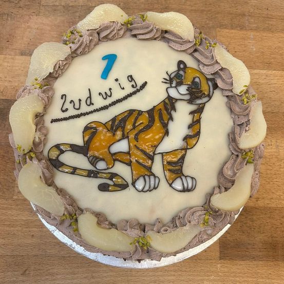 Ein Kuchen mit Tigerdesign und der Nummer 1 und dem Namen 'Zwdwig' darauf. Er hat Schokoladenglasur und ist mit kleinen Früchten verziert.