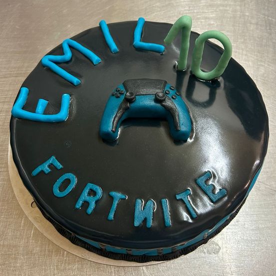 Ein schwarzer Geburtstagskuchen für Emilo, 10 Jahre alt, mit einem blauen Gamecontroller obenauf und Fortnite in Blau geschrieben.
