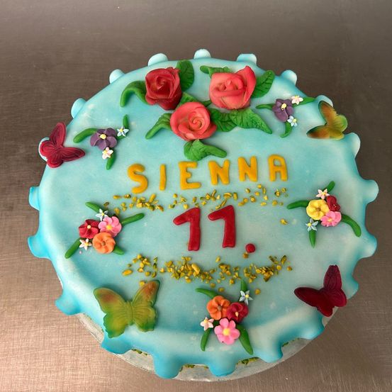Ein runder Kuchen mit blauer Zuckergussbasis, verziert mit roten Rosen, Schmetterlingen und Blumen. Es steht Sienna 11, mit gelben Blütenblättern auf dem Kuchen verstreut.