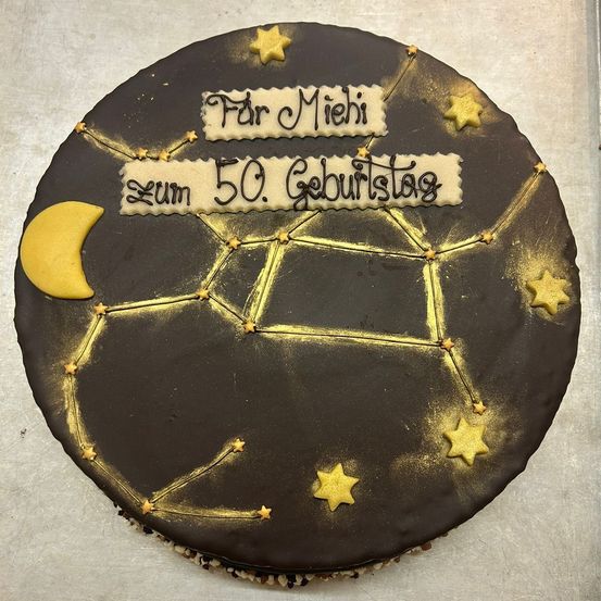Ein Kuchen aus dunkler Schokolade mit Sternbild und Sternen. Es hat einen Halbmond und steht 'Für Miehi Zum 50. Geburtstag'.