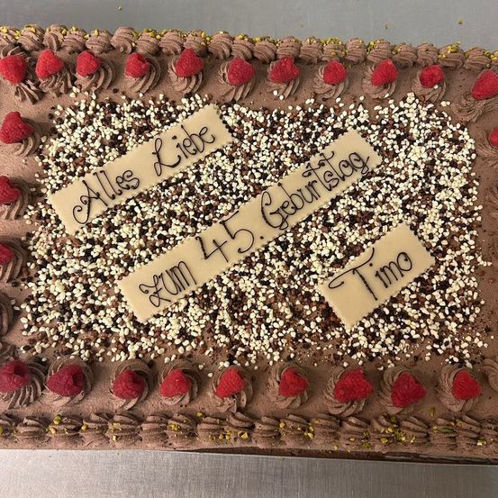 Ein Schokoladenkuchen mit weißen und braunen Streuseln, roten Himbeeren und weißer Glasur. Darauf steht 'Alles Liebe' und 'Zum 45. Geburtstag'. Auf einem separaten Stück Papier steht 'Timo'.