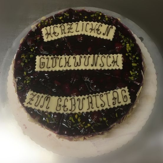 Ein Kuchen mit Blaubeerfüllung, überzogen mit einer Schokoladenglasur und Pistazien, mit der Aufschrift 'HERZLICHEN GLUCKWUNSCH ZUM GEBURTSTAG' darauf.