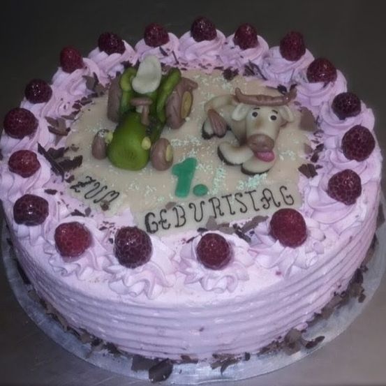 Ein Kuchen mit rosa Zuckerguss, geschmückt mit einem Traktor und einer Kuh, umgeben von Himbeeren und Schokolade. In der Mitte steht '1. Geburtstag'.