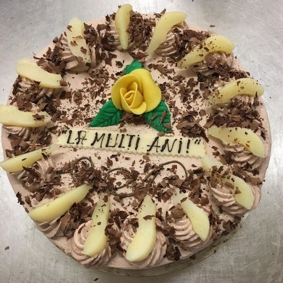Ein Schokoladenkuchen mit weißer Creme und weißen Fruchtstücken, verziert mit Schokoladenraspeln und einer gelben Rose. Die Worte 'La molti anni' sind darauf geschrieben.