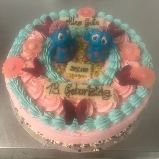 Ein Geburtstagskuchen mit rosa und blauer Glasur, der zwei blaue Monster mit Blumen und Schmetterlingen zeigt. Der Kuchen trägt den Text 'Alles Gute zum 13. Geburtstag'.