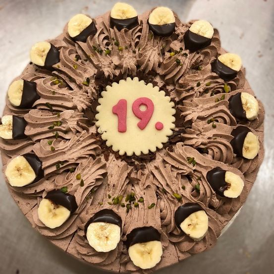 Ein Schokoladenkuchen mit Bananen- und Schokoladenstücken, verziert mit Schokolade und Pistazien, mit einer weißen kreisförmigen Nummer 19 in der Mitte.