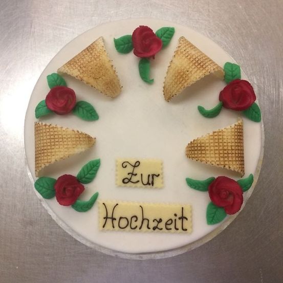 Ein Kuchen mit sechs Eiswaffeln, jede mit einer Rose und Blättern verziert. Der Kuchen hat 'Zur Hochzeit' auf Deutsch. Er steht auf einer metallischen Oberfläche.