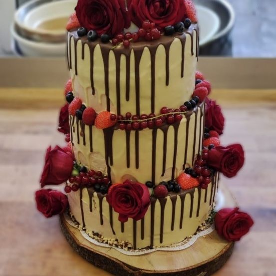 Dreistufiger Kuchen mit gelber Glasur, roten Rosen und Beeren. Schokoladen-Drizzles schmücken die Seiten.