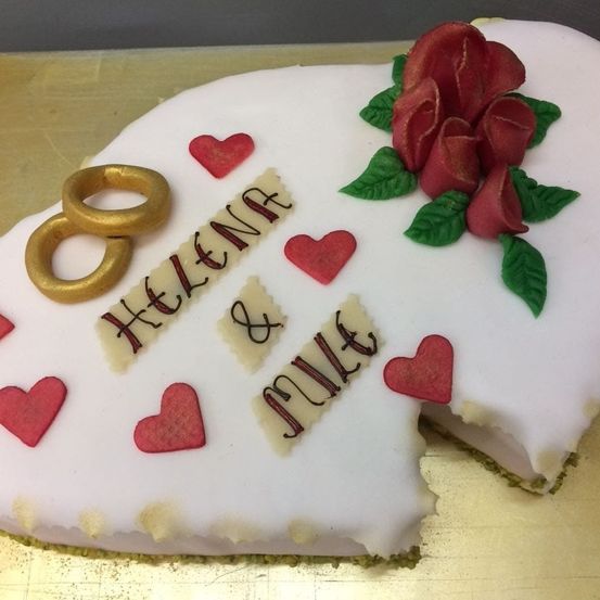 Ein herzförmiger Kuchen mit 'Helena & Julia' in Zuckerguss geschrieben, verziert mit roten Herzen und goldenen Eheringen, gekrönt von einer Rose und grünen Blättern.