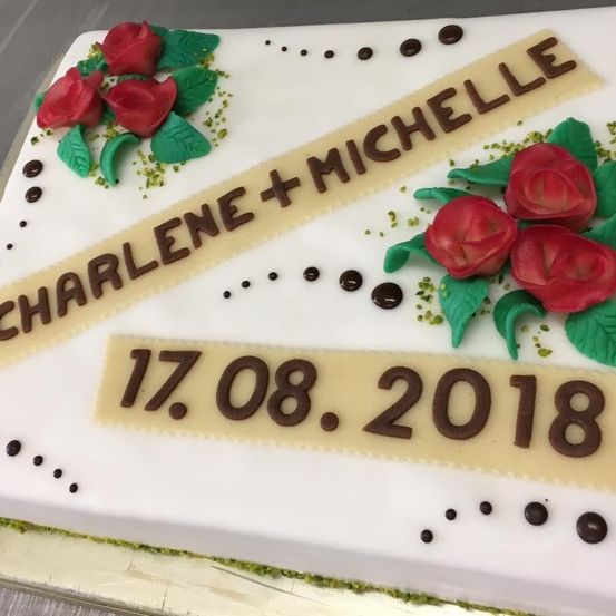 Ein weißer Kuchen mit roten Rosen und grünen Blättern, die Namen Charlene und Michelle zeigen. Darunter steht das Datum 17.08.2018 in Schokolade geschrieben.