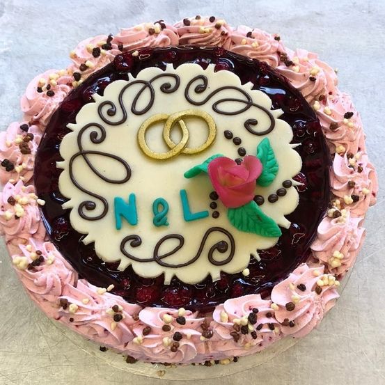 Ein runder Kuchen mit rosa Zuckerguss, Schokostückchen und schwarzer Kirschfüllung. Er hat zwei goldene Ringe und die Buchstaben N&L mit einer rosa Rose obenauf.