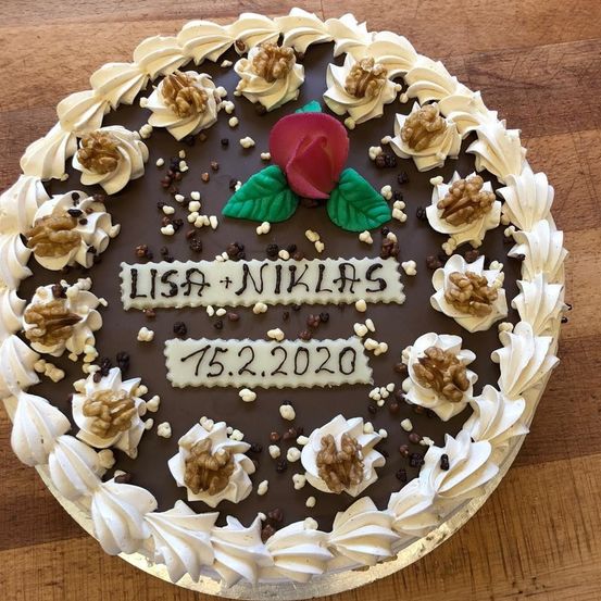 Ein Schokoladenkuchen mit weißer Glasur, Walnüssen und einer roten Rose mit grünen Blättern, dekoriert mit 'LISA + NIKLAS' und '15.2.2020' auf einem Holztisch.