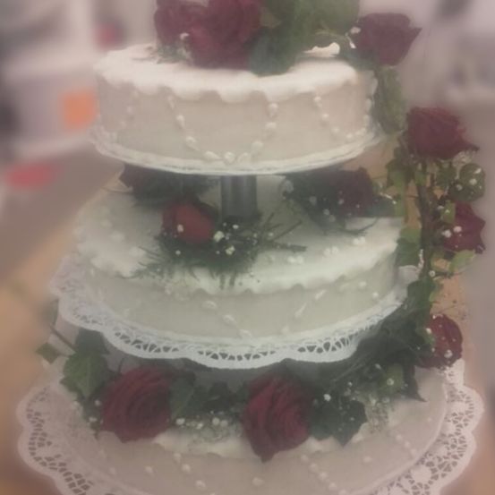 Ein dreistufiger Hochzeitskuchen mit weißer Glasur, roten Rosen und grünen Blättern, der auf einer Spitzenserviette ruht.