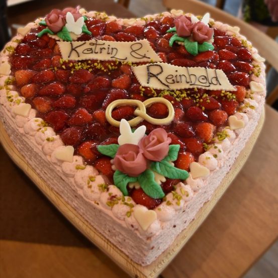 Ein herzförmiger Kuchen, verziert mit Erdbeeren, weißer Glasur und Blumen. Es sind zwei Namen, Katrin und Reinhold, darauf geschrieben. Oben befinden sich auch zwei goldene Ringe.