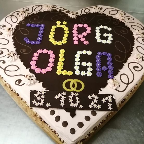 Ein herzförmiger Kuchen, verziert mit bunten Buchstaben, die 'JORGE OLGA' und gelbe Eheringe bilden, umgeben von weißen und schwarzen Punkten.
